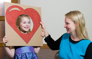 Little girl with heart cutout around her face held up by a teacher