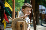 A little boy stands at the ships wheel on the helm in a playground setting. He's pretending to navigate his ship through the waters.
