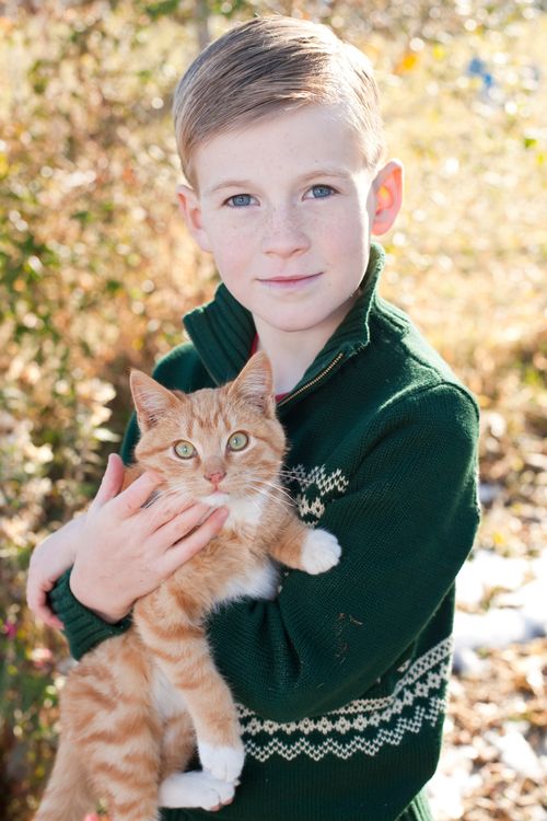 A young boy with blonde hair and a green sweater standing and holding a light orange cat in his arms.