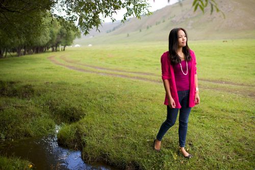 A young Mongolian woman wearing a pink sweater stands next to a stream in a green field, looking into the distance.