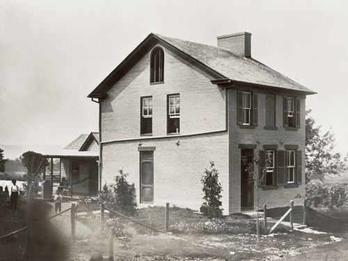 Black and white photo of a two-story home built of light brick with an annex in the back.