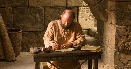Attēlā vīrietis pie rakstāmgalda raksta uz pergamenta