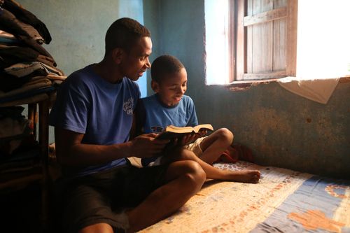 A father sitting on a bed, reading scriptures with his young son by the light from a window in their home in Madagascar.