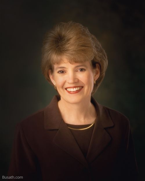 A photograph of Susan Winder Tanner against a dark gray background, wearing a dark brown blazer and gold necklace.