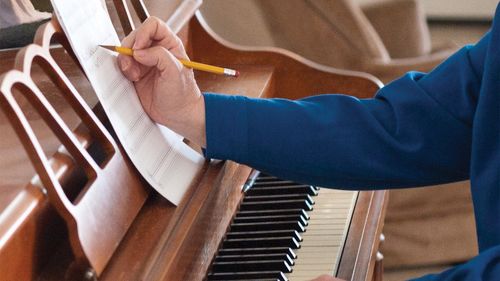A woman sits at a piano and touches the keys with one hand and makes notes on her sheet of music with the other.