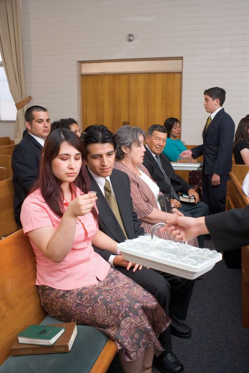 A woman in a pink blouse sits near her husband in a chapel and takes a cup of water from a sacrament tray.