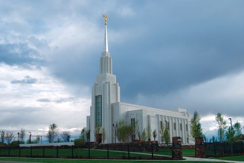 The front and side of the Twin Falls Idaho Temple behind a black fence on a cloudy day.
