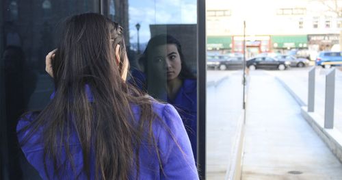 A woman is standing looking at her refection in the widow of a building. You can see a city street in the background.