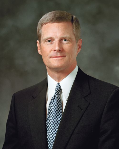 A portrait of Elder David A. Bednar, who is wearing a black suit with a black and white geometric tie, in front of a gray background.