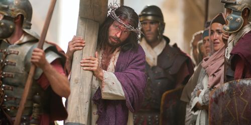 Jesus carries His cross on the way to His Crucifixion while wearing a crown of thorns and a purple robe placed on Him by soldiers.