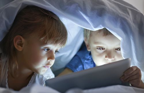 two children look at a tablet while hiding underneath a blanket
