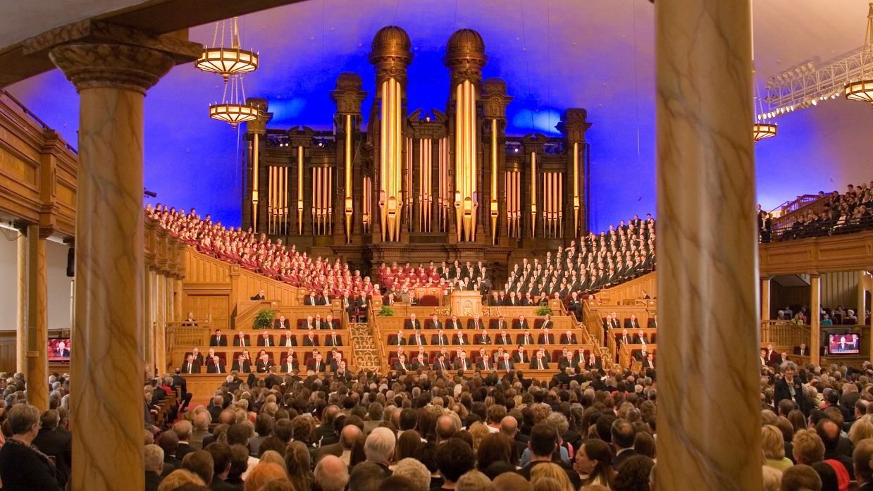 A view from the back of the Salt Lake Tabernacle, showing two pillars and many rows of benches filled with people.