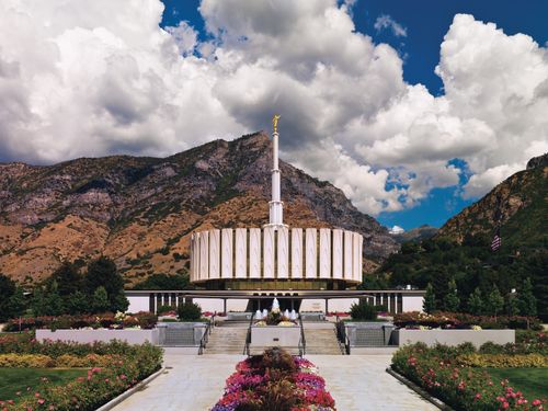 A wide-angle view of the Provo Utah Temple, with large mountains in the background and white clouds overhead.