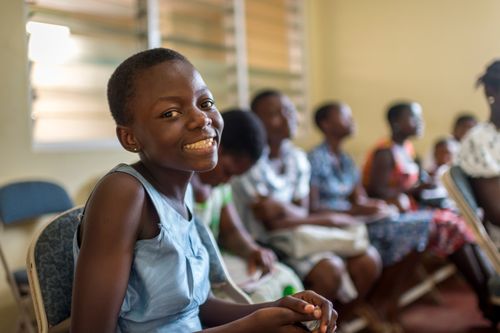 A young girl in a blue dress sits in Sunday School with other youth and smiles.