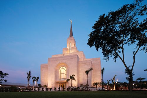 The entire San Salvador El Salvador Temple in the evening, with windows lit from inside and the temple lit on the outside, surrounded by trees on the temple grounds.