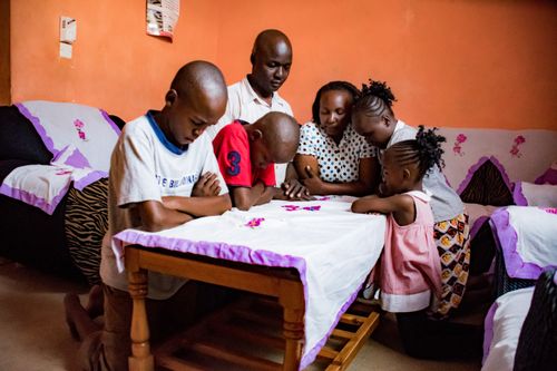 A family of four children and parents in Mombasa, Kenya, kneeling in prayer around a table in their home.