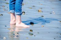 Bare feet of girl, standing on beach with reflections and small rocks.