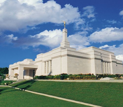 A front and side view of the Mérida Mexico Temple, with green grass and a blue sky with clouds.