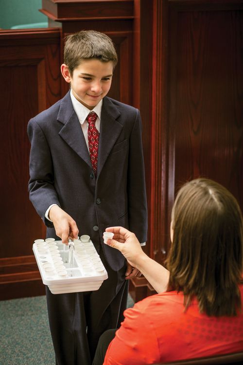 A young man in a suit holds out the sacrament water tray to a woman in a red blouse.