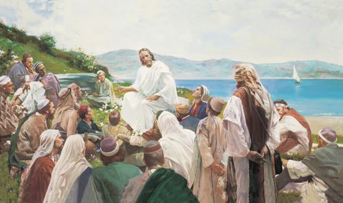 Jesus in white robes, sitting on a hillside by the sea, surrounded by a large group of people who are listening to His teachings.