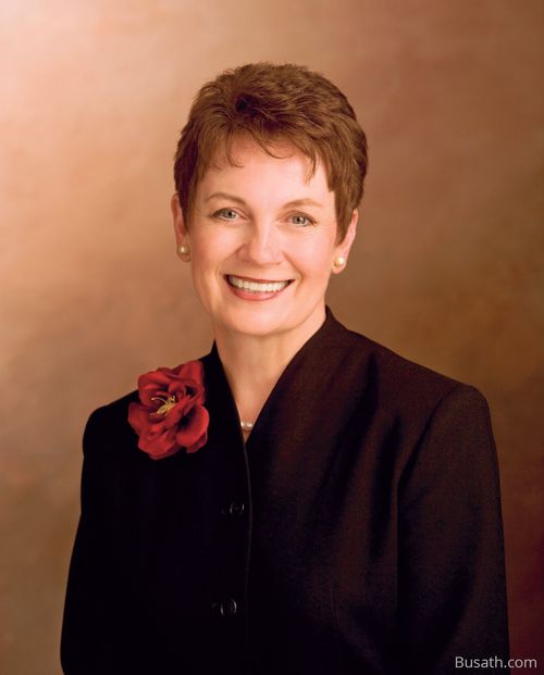 A photograph of Cheryl C. Lant against a brown background, wearing a black blazer with a red rose pin.