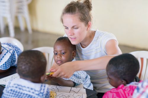 A woman sits in a chair and helps three small children color at a table in an orphanage in Ghana.