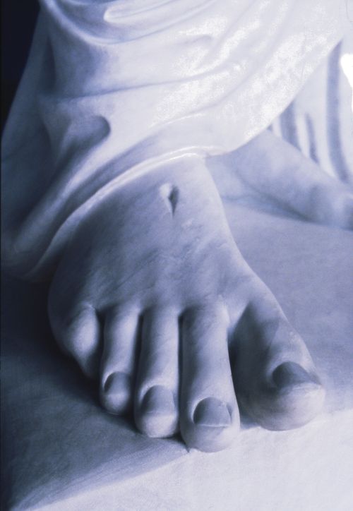 A close-up photograph of the foot of a white statue of Jesus Christ, showing the nail print and the hems of Christ’s robes.
