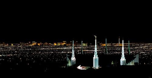 A far-off view of the Las Vegas Nevada Temple spires, illuminated with white light at night, with the city lights in the distance.