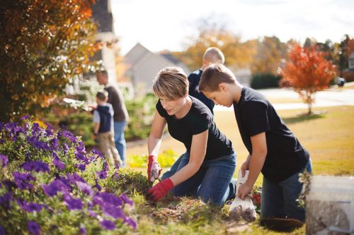 A mother and son wearing black t-shirts and blue jeans while working in a flower bed on a sunny day with the rest of the family in the background.
