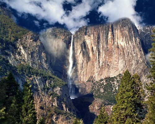 A large waterfall over a cliff bordered by trees and a blue sky in Yosemite National Park.