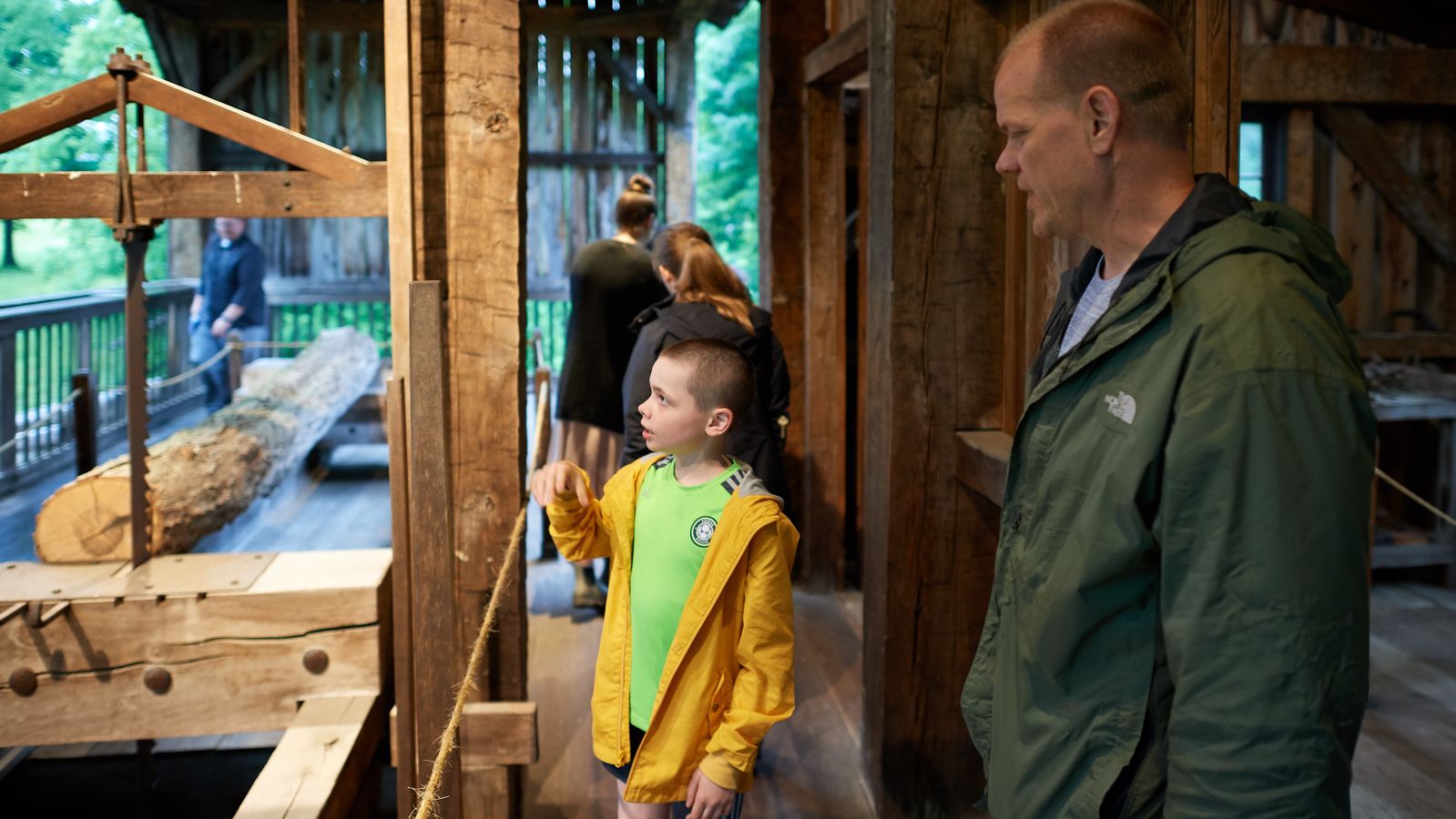 Standing inside a sawmill, a man and young boy look at a vertical saw in a wooden frame.  A long log is positioned in front of the saw’s teeth.  