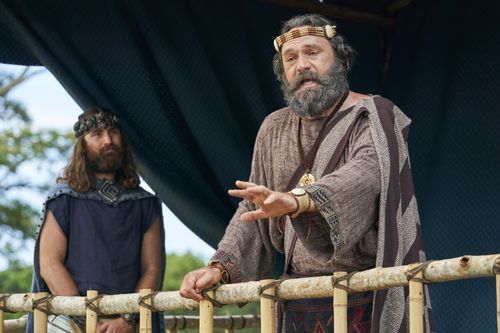 King Benjamin stands on a tower speaking to the Nephites and Mosiah watches in the Land of Zarahemla.