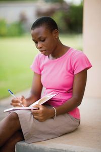 A young woman in Ghana sits outside on a step and writes in a journal.