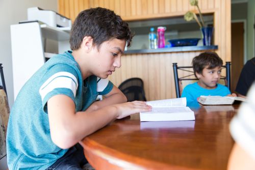 A teenage boy sitting at a table studying the scriptures while his brother sits nearby 