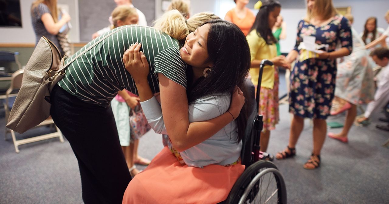 Woman hugging a young woman in a wheelchair. Other young women and leaders in the background. They are inside an LDS Chapel.