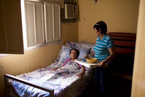 A woman in Paraguay bringing a tray of food to an elderly woman sick in bed.