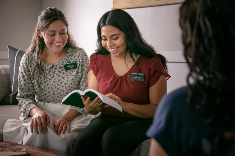 Two sister missionaries study the Book of Mormon with a woman on her living room couch