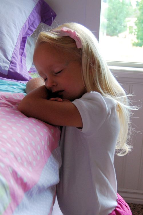 A girl wearing a white T-shirt and a pink bow in her blonde hair kneels next to her bed with her eyes closed to say a prayer in the morning.
