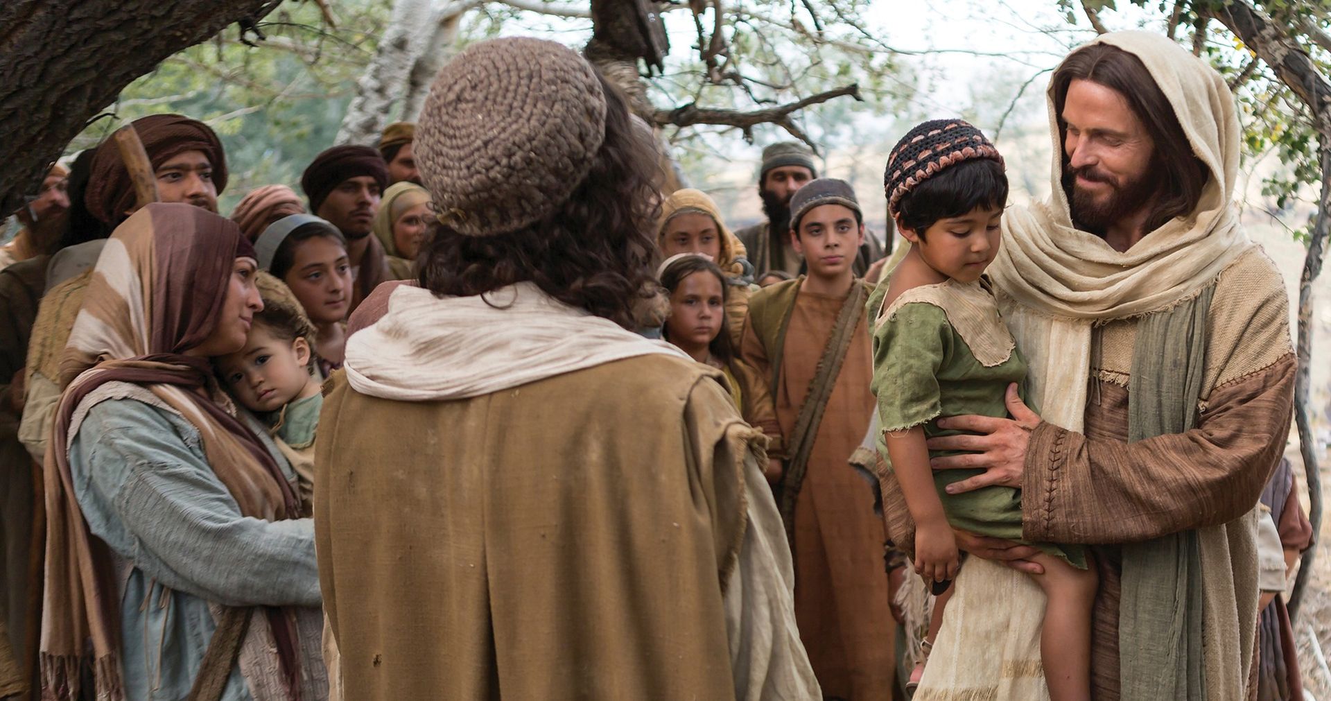 Jesus is sitting down on the ground with a young boy in his lap and surrounded by mothers and other people.  Outtakes include Jesus holding children and walking with them, his disciples with him, Jesus and Peter, Jesus with Peter and James and some of the children. Still image from The Life of Christ Bible Videos: The Gospels video.
