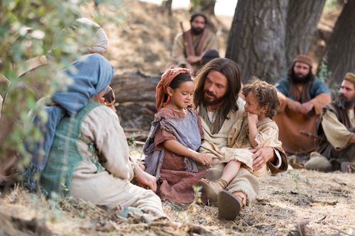 Jesus is sitting down on the ground with a young child in His lap, surrounded by mothers and other people.