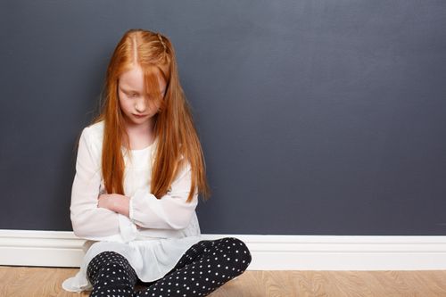 A young girl with red hair sitting on the floor with her head bowed and arms folded in prayer.