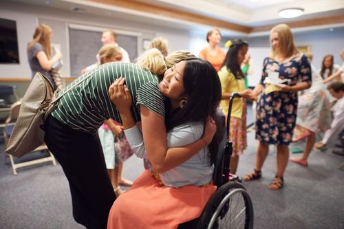 A young woman in a green striped shirt and black skirt leans down to hug her friend who is sitting in a wheelchair.