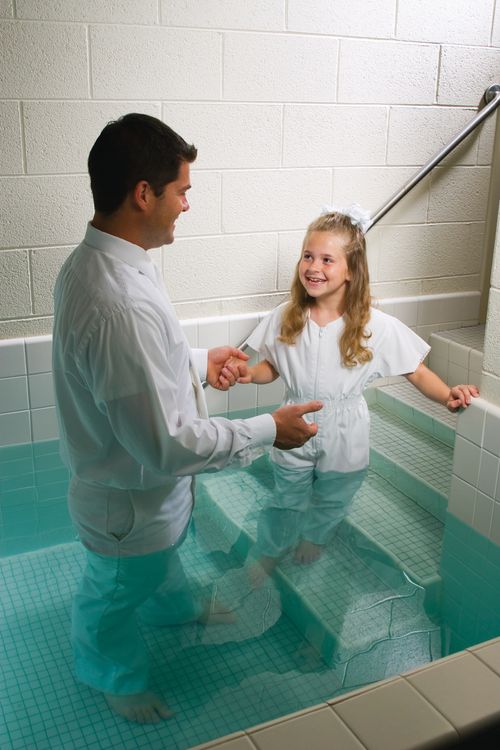 A young girl in a white jumpsuit walks toward her father, who is standing in a baptismal font.