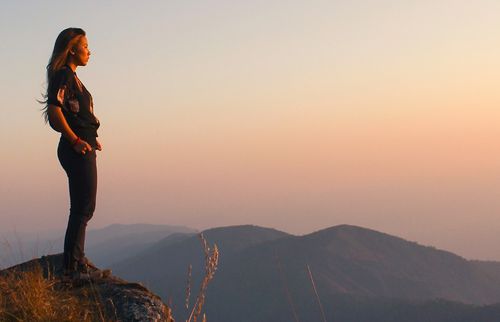 a woman standing on a mountain ledge looking at the sunset