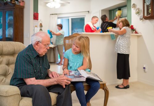 A little girl sits on a chair with a book in her lap and reads it to her grandfather, who sits in a chair next to her, while other family members clean the kitchen in the background.