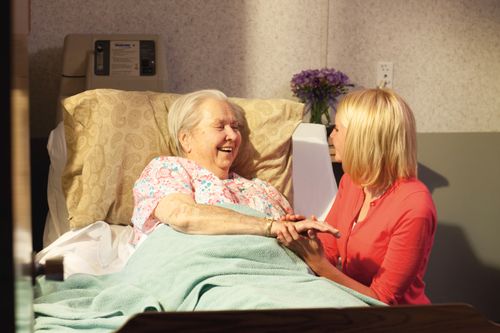 A young woman visits and holds the hand of an elderly woman lying in a hospital bed.