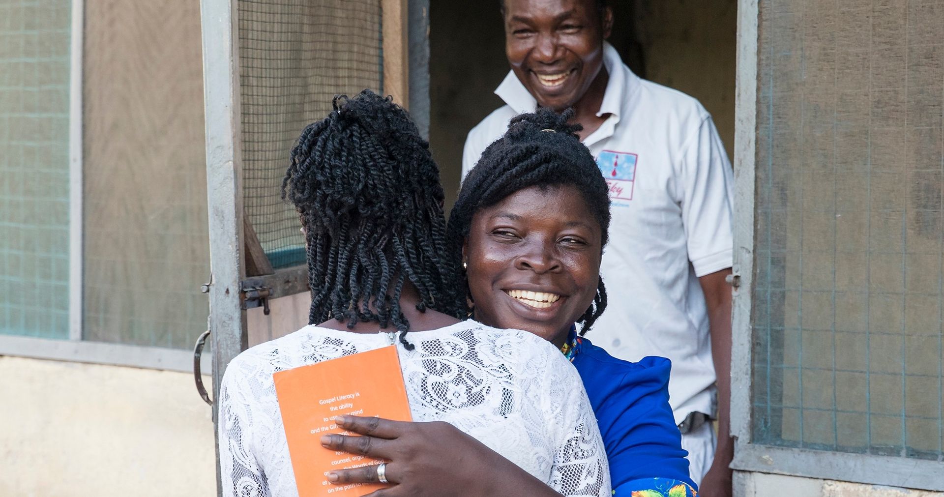 Two Ghanaian women embrace. A Ghanaian man smiles in the background.
