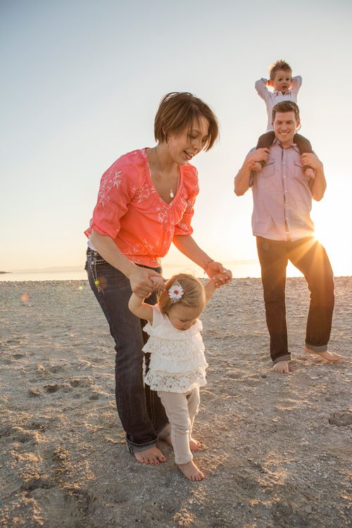 A mother holds her baby daughter’s hands while walking on the beach with her husband and other child.