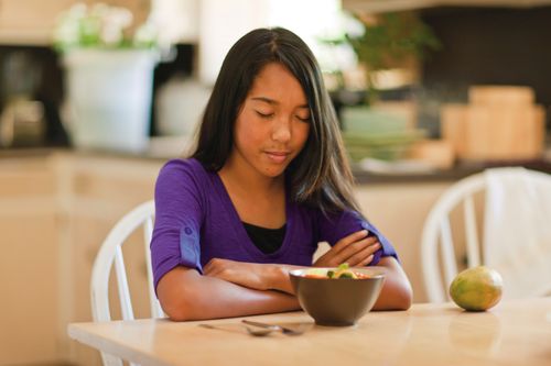 A young girl folds her arms and prays while sitting at a dinner table, about to eat.