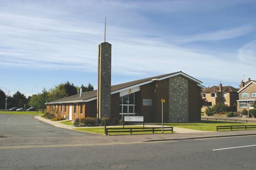 The outside of a dark brown chapel in Ireland surrounded by green grass and a fence in front with a parking lot on the side.